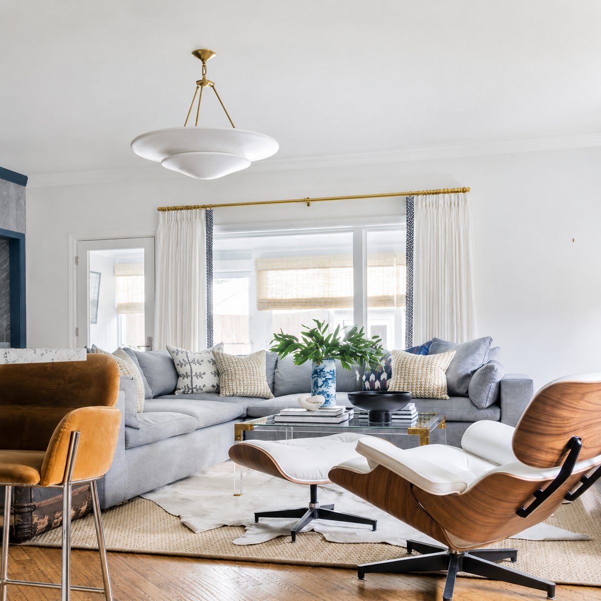A modern living room featuring a light gray U-shaped sectional sofa with patterned throw pillows, a mid-century style wooden lounge chair with a white cushion, a matching ottoman, a glass coffee table, and a large window with white curtains and gold trim.