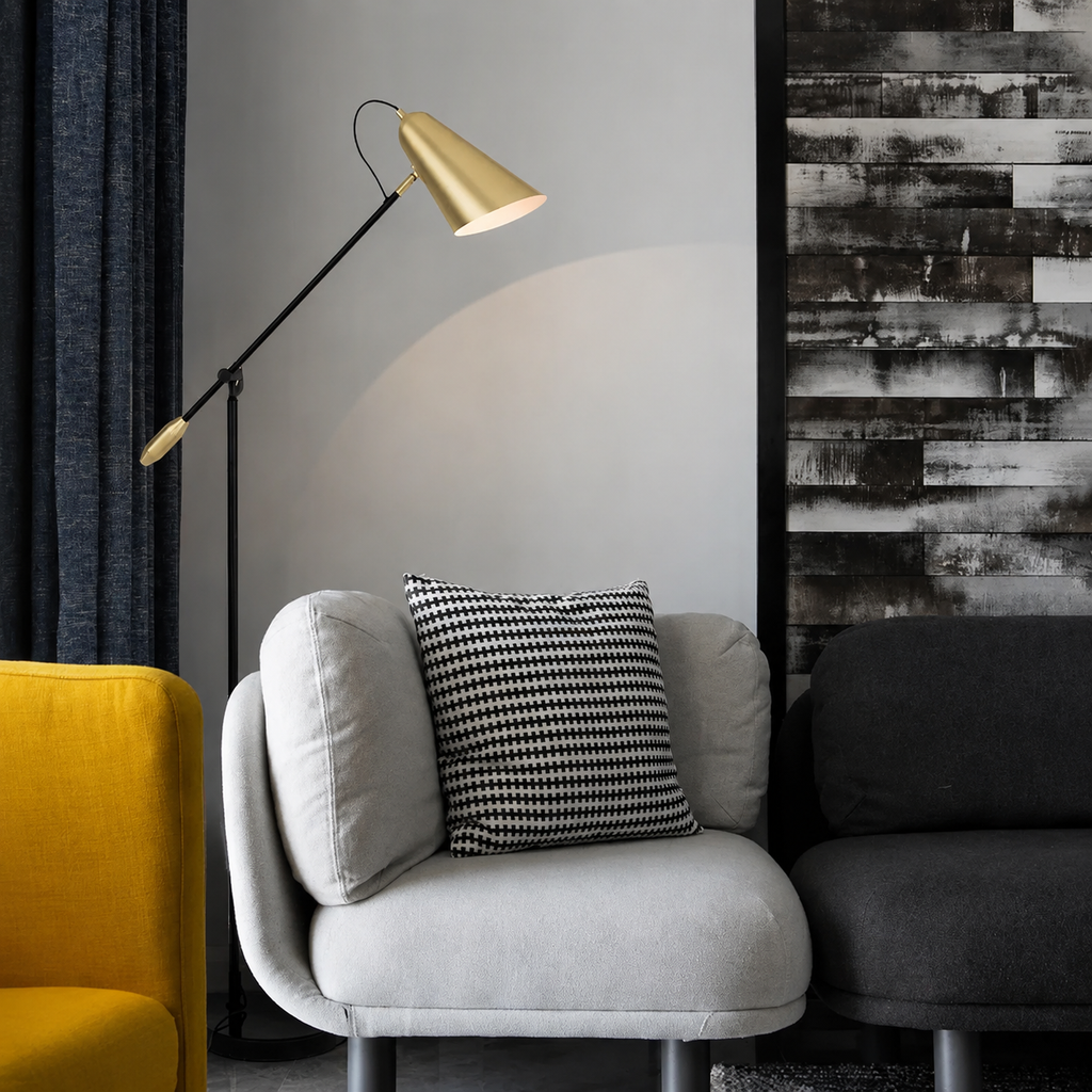 A modern black floor lamp with a gold metal cone shade stands next to a light gray upholstered armchair with a black-and-white patterned throw pillow. The scene is set in a styled interior with a dark gray sofa, a yellow chair edge, and a textured black-and-white wood-paneled wall.