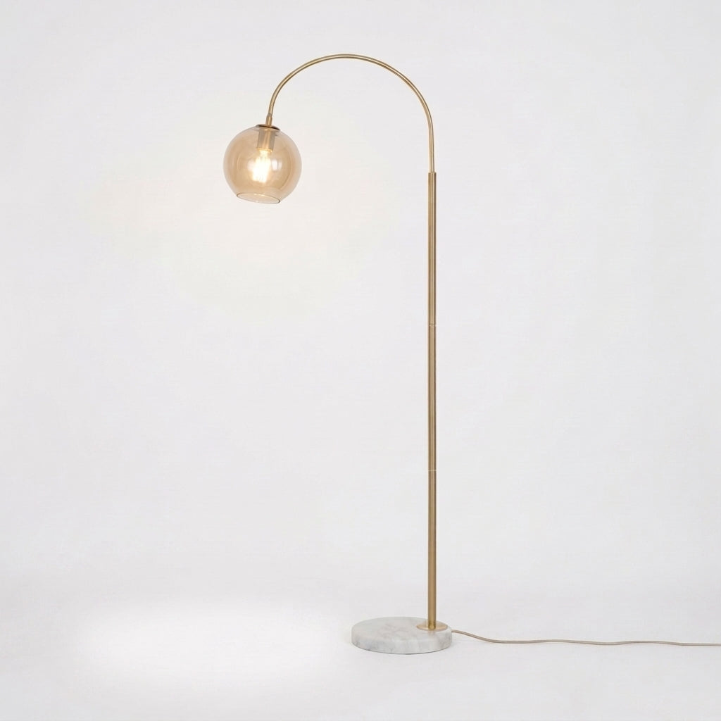 A modern floor lamp with a curved brass-colored metal arm, a spherical glass shade, and a round white marble base, shown lit against a plain white background.
