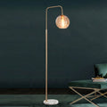 A modern floor lamp with a slim gold-finished metal stand, a curved top arm, a clear glass globe shade, and a round white marble base, shown lit in a styled interior next to a green upholstered chair.