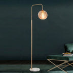 A modern floor lamp with a slim gold-finished metal stand, a curved top arm, a clear glass globe shade, and a round white marble base, shown lit in a styled interior next to a green upholstered chair.