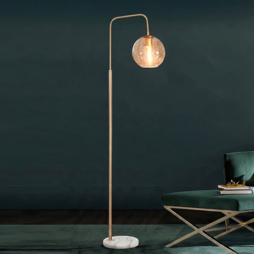 A modern floor lamp with a slim gold-finished metal stand, a curved top arm, a clear glass globe shade, and a round white marble base, shown lit in a styled interior next to a green upholstered chair.