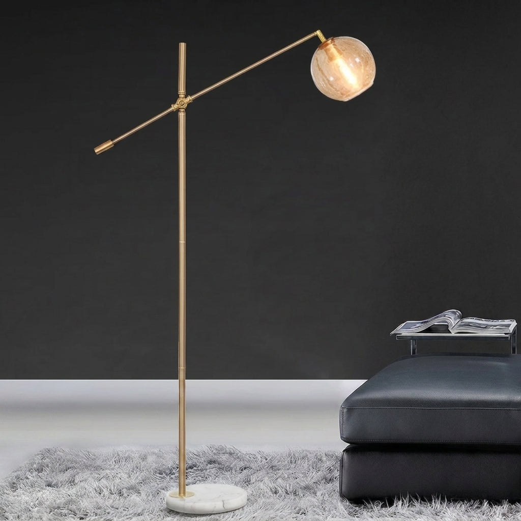 A modern floor lamp with a slim gold-finished metal stand, an adjustable arm, and a round glass shade, shown in a styled interior setting next to a dark leather ottoman.
