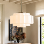 A modern multi-tiered pendant light with a frosted white glass or acrylic shade and a brushed brass or gold-toned metal top canopy and rod, hanging in a styled interior room with a window, artwork, and decor.