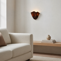 A modern sculptural wall sconce with a dark amber-brown fluted glass shade, mounted on a warm off-white wall in a minimalist living room setting next to a cream-colored sofa.