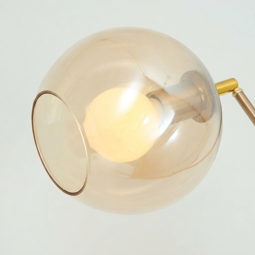 A modern spherical wall lamp with a warm amber-tinted glass shade, glowing Edison bulb inside, and a brass metal arm against a light gray background.