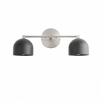 A modern two-light wall sconce with matte black dome-shaped shades and a brushed nickel circular backplate and horizontal arm, shown against a plain white background.