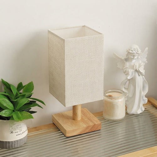 A table lamp with a textured beige linen rectangular shade and a light natural wood base, styled on a ribbed glass tray next to a small green plant and a white angel figurine.