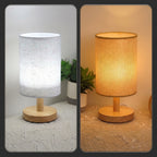 A split-screen view of a table lamp showing two light modes: on the left, the lamp glows with cool white light from a textured white fabric shade and a light wood base; on the right, it glows with warm yellow light under the same shade and base. Both are shown on a patterned beige tablecloth with a small potted plant in the background.
