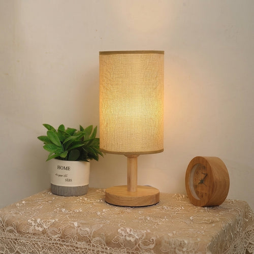 A warm-toned table lamp with a cylindrical linen-textured shade and a round wooden base sits on a lace-covered table. A small potted green plant and a round wooden clock are placed beside it against a plain wall.
