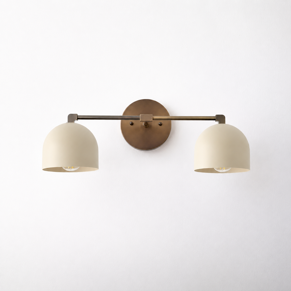 A modern two-light wall sconce with dome-shaped cream shades and a warm bronze circular backplate, mounted on a plain white wall.