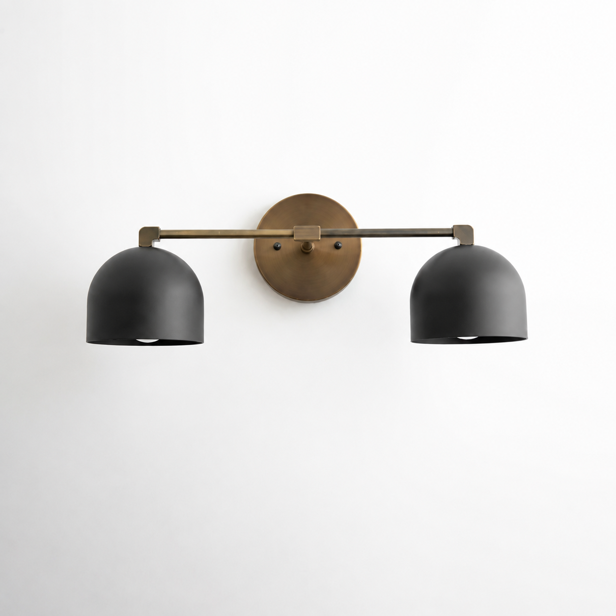 A modern two-light wall sconce with matte black dome-shaped shades and a brushed brass-finished horizontal arm and circular wall mount, shown against a plain white wall.